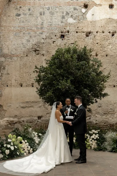 Wedding vows as bride in cathedral veil and groom in black tux hold hands while officiant reads into a microphone in a courtyard