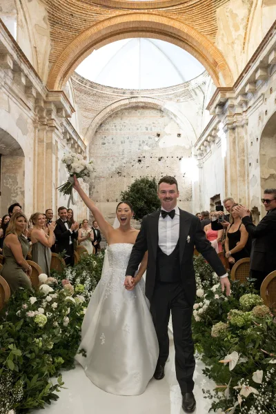 Wedding recessional as newlyweds walk hand in hand down a greenery-lined aisle, bride raising bouquet beneath arched stone ceiling and cheering guests