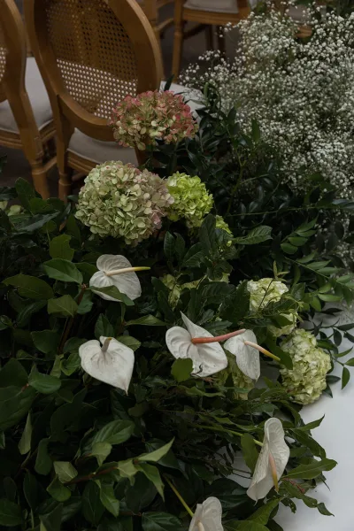 Wedding aisle flowers with hydrangeas and white anthurium lining a white runner, grounded meadow-style florals beside wooden chairs indoors