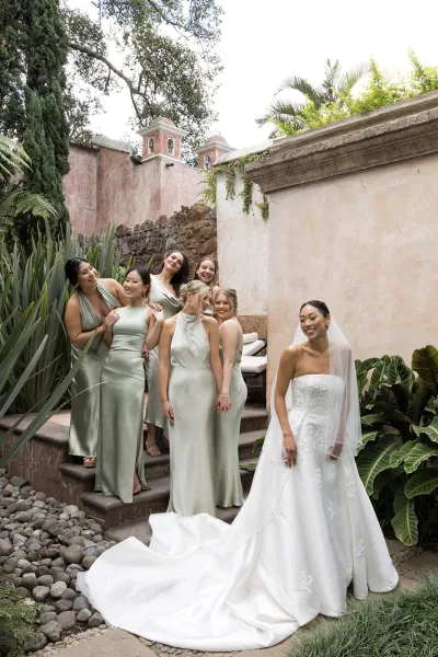 Bride with bridesmaids in sage green dresses on stone steps, bride in strapless lace gown with long train and veil, tropical plants behind