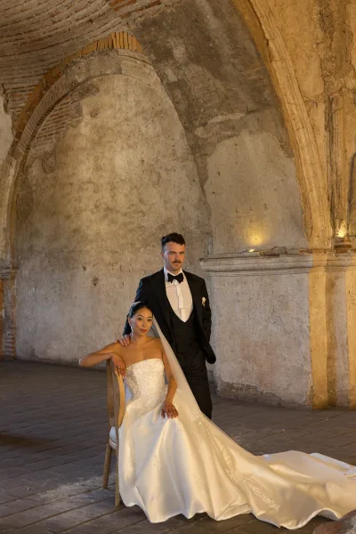 Couple portrait with bride seated in a strapless lace wedding dress and veil, groom in black tuxedo beneath warm-lit stone arches