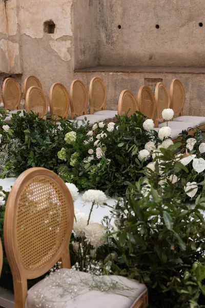 Ceremony aisle decor with ground floral aisle of white blooms and lush greenery, flanked by rattan bistro chairs in a stone-walled venue