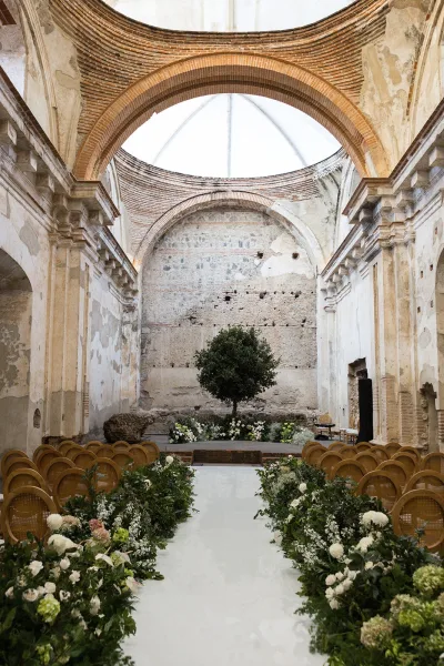 Ceremony setup with white aisle runner and greenery garland, wooden chairs facing a tree altar inside stone ruins with brick arches