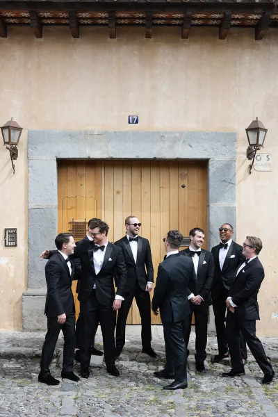 Groomsmen portrait in matching black tuxedos and bow ties, one wearing sunglasses, posed by a stone doorway on a cobblestone street