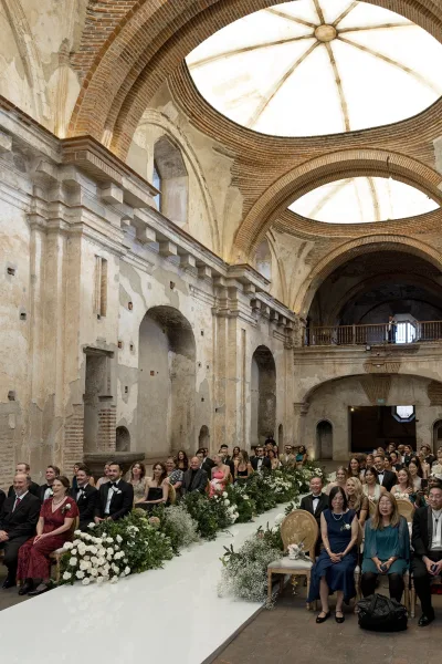 Ceremony setup with a white aisle runner and greenery-lined aisle, white roses and baby's breath in a historic stone interior with brick arches