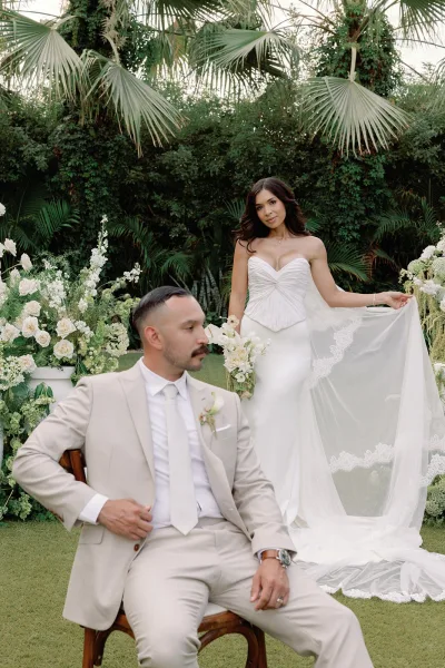 Couple portrait with bride holding veil beside her seated groom, framed by palm trees and lush greenery on a manicured lawn.