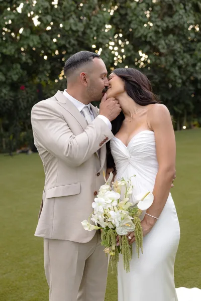 Wedding kiss portrait of the bride and groom kissing on a garden lawn, the bride holding a modern white anthurium orchid bouquet.