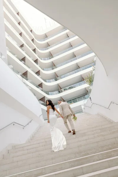 Couple portrait of bride and groom walking hand in hand on glass-railed modern stairs, as he carries the bouquet against white walls and balconies.