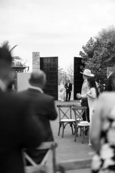 Ceremony processional with a bride walking down the aisle, emerging through large wooden doors into a palm-lined courtyard.
