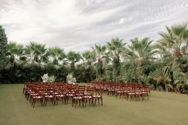 Outdoor ceremony setup with garden ceremony seating in wooden crossback chairs, framed by palm trees and tropical greenery on a sunlit lawn.