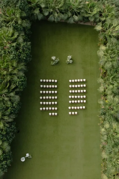 Ceremony setup for an outdoor wedding ceremony with floral arrangements on a grass lawn, framed by palm trees and garden hedges.