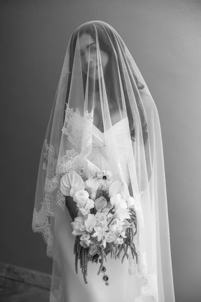 Bridal portrait of a bride under a veil, softly posed with a bouquet adorned with photo locket charms.