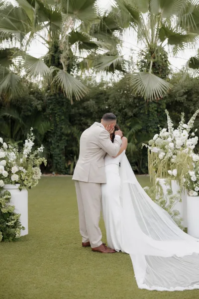 Ceremony kiss moment as the bride and groom share their wedding first kiss beneath palm trees, her long veil trailing on the garden lawn.