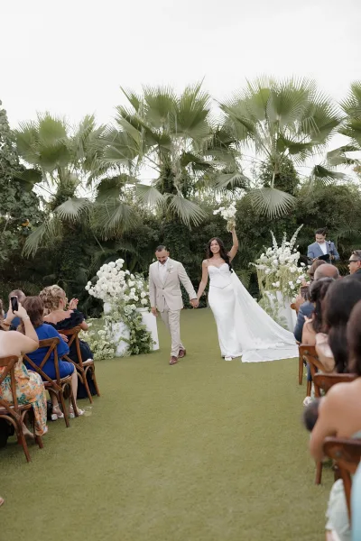 Wedding recessional with a just married exit as the bride and groom walk the aisle, the bride lifting a white bouquet amid palm trees and guests.