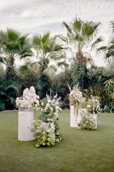 Wedding altar showcasing outdoor ceremony decor adorned with white florals, framed by palm trees and tropical foliage on a grass lawn.