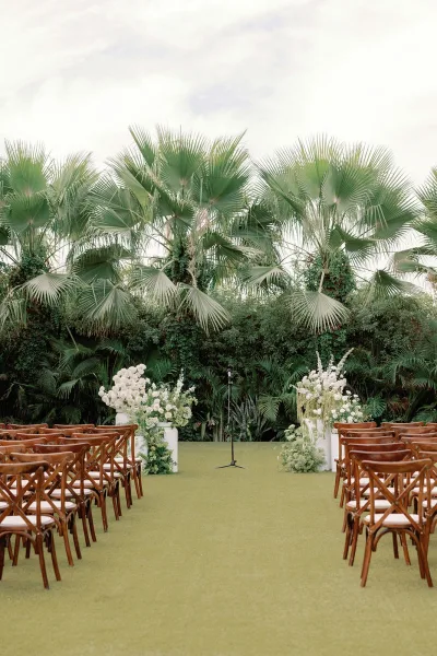 ceremony setup featuring a clean ceremony aisle design with wood crossback chairs, framed by palm trees on a sunny garden lawn