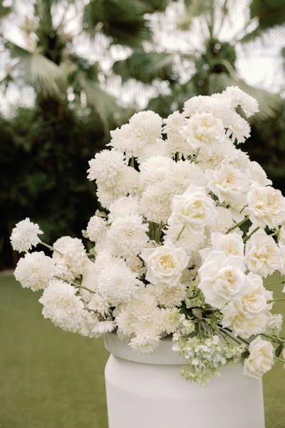 Wedding floral arrangement featuring a white rose arrangement displayed in a pedestal vase on a garden lawn framed by palm trees.