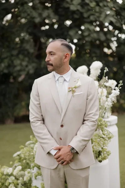 Groom portrait of a beige suit groom with a single boutonniere, in a garden with trees and white pedestal vases.