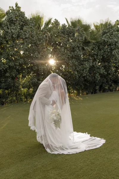 Couple portrait of a bride and groom under a flowing veil, sharing a tender moment on a sunlit lawn framed by palm trees and a leafy hedge.