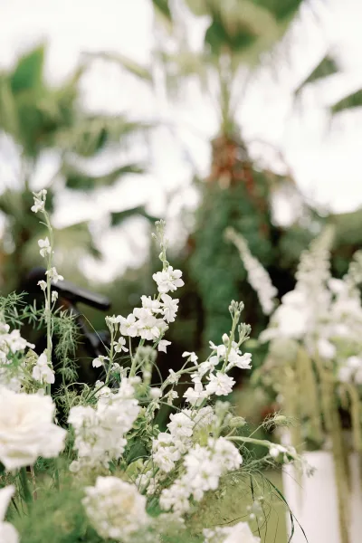 Ceremony flowers in a white floral arrangement atop a pedestal, framed by palm trees, garden greenery, and an open sky