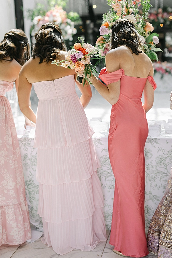 Bridesmaid dresses in mismatched blush and coral tones, with one holding a bouquet beside reception tables with bokeh lights