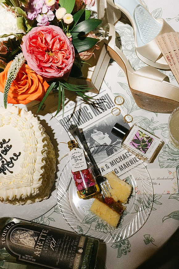 Wedding flat lay of cake slices, wedding rings, heels, perfume, champagne, place card and bouquet on a patterned floral tablecloth