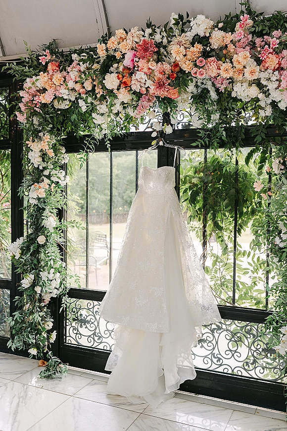 Wedding dress hanging on a hanger, a strapless lace gown with tulle skirt framed by a pink and peach floral arch in a conservatory