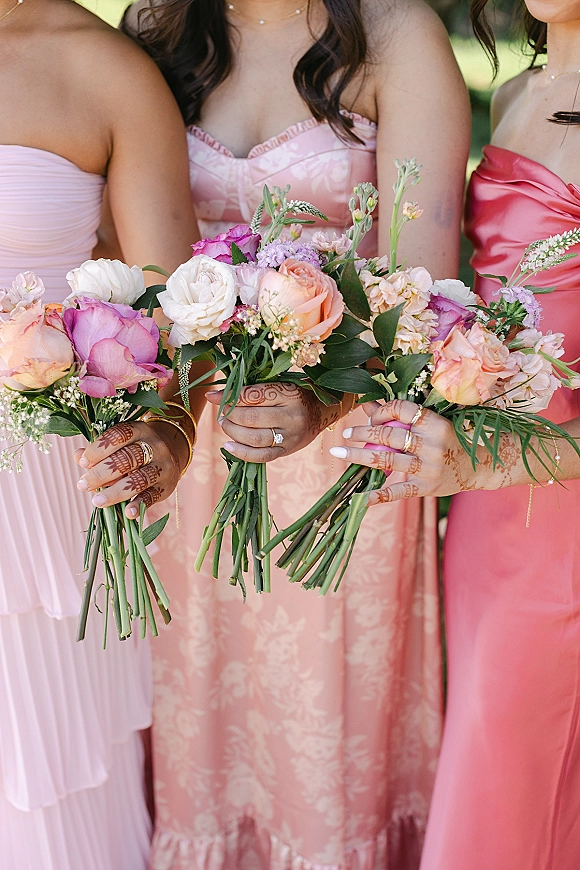 Bridesmaid bouquets of pink and peach bouquets with rose blooms and greenery, held by bridesmaids with henna and rings outdoors