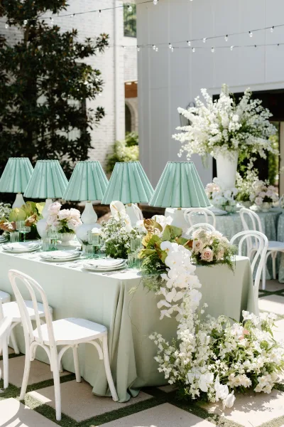 Reception tablescape with sage green linens, mint pleated lamps, orchids and blush roses on an outdoor patio under string lights