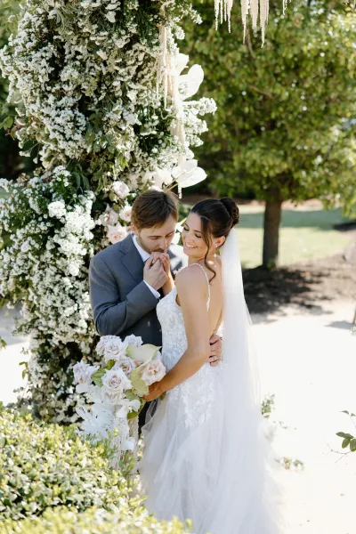 Couple portrait of groom kissing bride’s hand as she holds a blush rose bouquet beneath a white floral arch in a sunlit garden