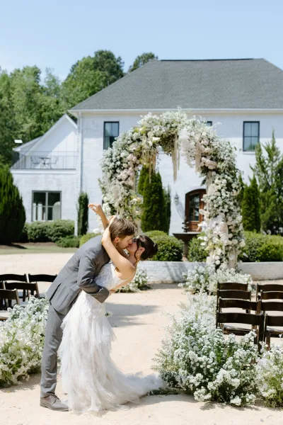 Wedding kiss portrait of bride in lace gown and groom in gray suit dipping beneath a white floral arch on an outdoor lawn by a house