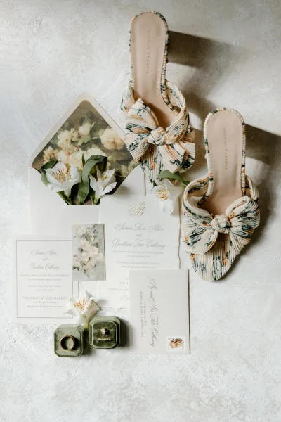 Invitation suite wedding invitation flat lay with watercolor card, calligraphy envelopes, RSVP, rings, bow heels, and white blooms on textured surface