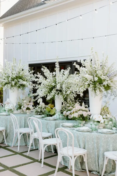 Reception tablescape with tall urn floral centerpieces, blush roses and greenery, pale blue linens, green goblets, and string lights by a white barn patio