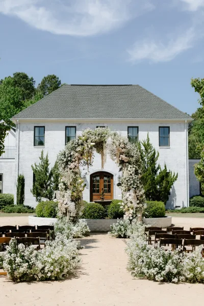 Ceremony arch decor with white flowers and greenery, hanging strands and aisle florals set in a gravel courtyard by a white brick building