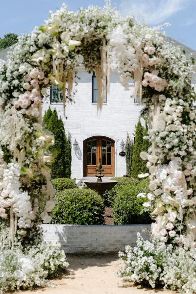 Wedding floral arch with white flowers and blush roses draped in greenery, framing a garden fountain before white brick doors under blue sky