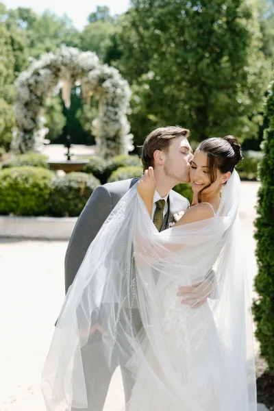 Wedding couple portrait of groom kissing bride’s cheek as she looks over her shoulder, cathedral veil draped by a floral arch in a garden walkway