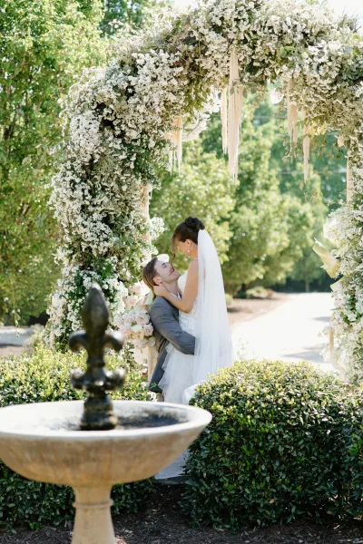 Wedding couple portrait of bride and groom embracing beneath a floral arch, her veil and bouquet catching sunlight near a garden fountain