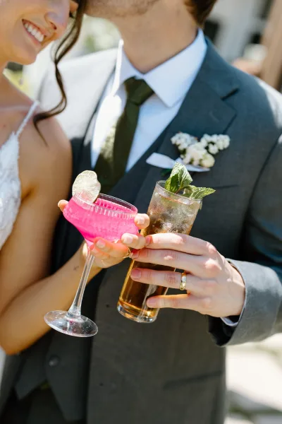Wedding cocktail toast as bride and groom cheers with pink coupe and mojito highball, mint and lime garnish, blurred outdoor guests behind