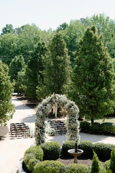 Ceremony setup for an outdoor wedding ceremony with a floral arch of white flowers and hanging strands, wooden chairs along a gravel aisle in a garden