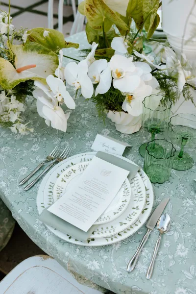 Reception tablescape with wedding place setting, white orchid centerpiece and anthurium leaves, green goblets on a patterned cloth outdoors