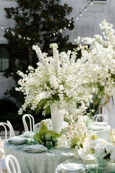 Reception tablescape with wedding table decor featuring tall white floral centerpiece, green goblets, patterned cloth, string lights on patio