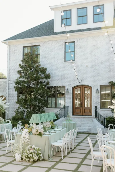 Reception tablescape with sage green linens, white bentwood chairs, floral garland and green glassware under string lights on a patio by a white brick building