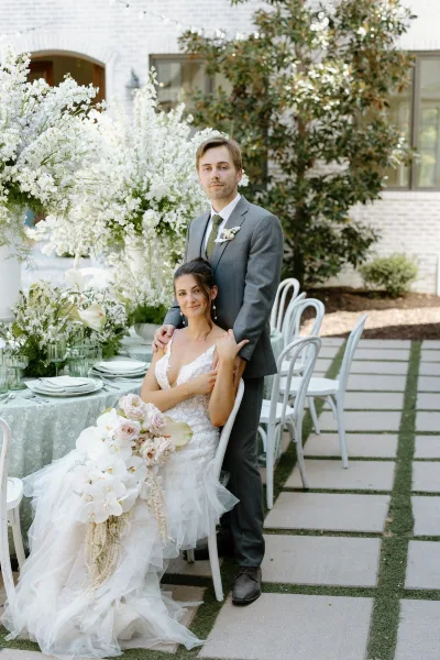 Couple portrait of bride in lace dress holding a cascading orchid bouquet as groom in gray suit stands behind at a garden courtyard table setting