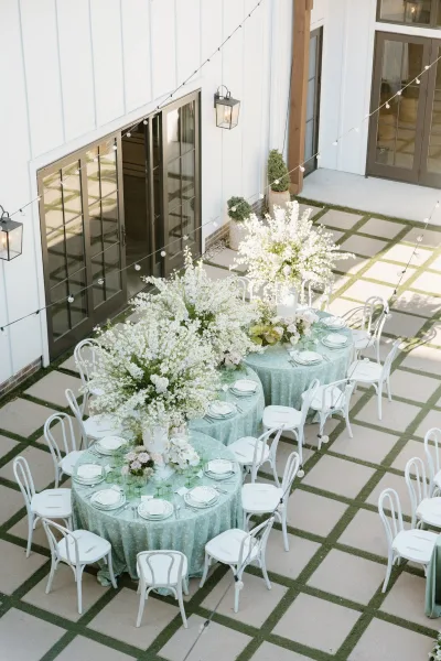 Reception tablescape with outdoor reception tables dressed in seafoam linens, tall white florals, and green goblets under string lights in a barn courtyard patio