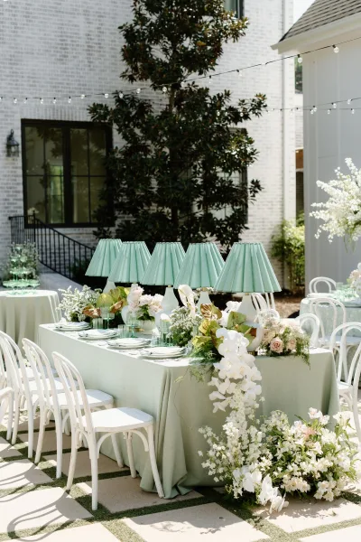 Reception tablescape with mint green linen, pleated green lamps, orchids and blush roses, green glassware under patio string lights by brick windows