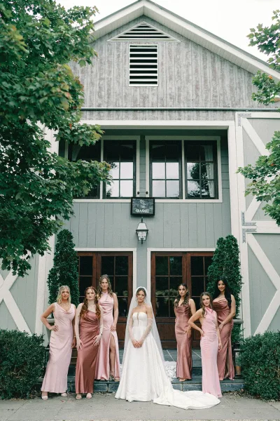 Bridal party portrait of the bride with bridesmaids in mismatched mauve dresses, framed by a barn facade and greenery with a veil accent