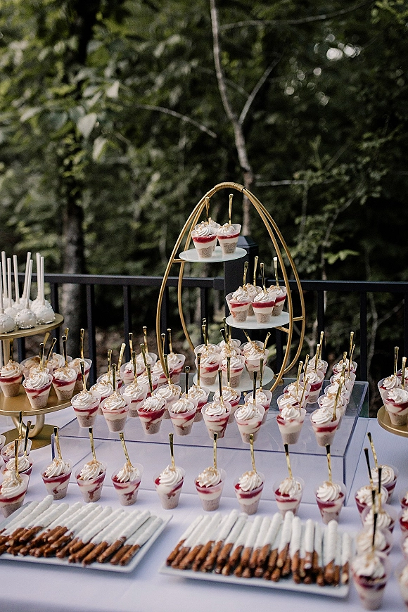 Dessert table with mini dessert cups topped with whipped cream, gold dessert spoons, and pretzel rods on a white cloth by trees