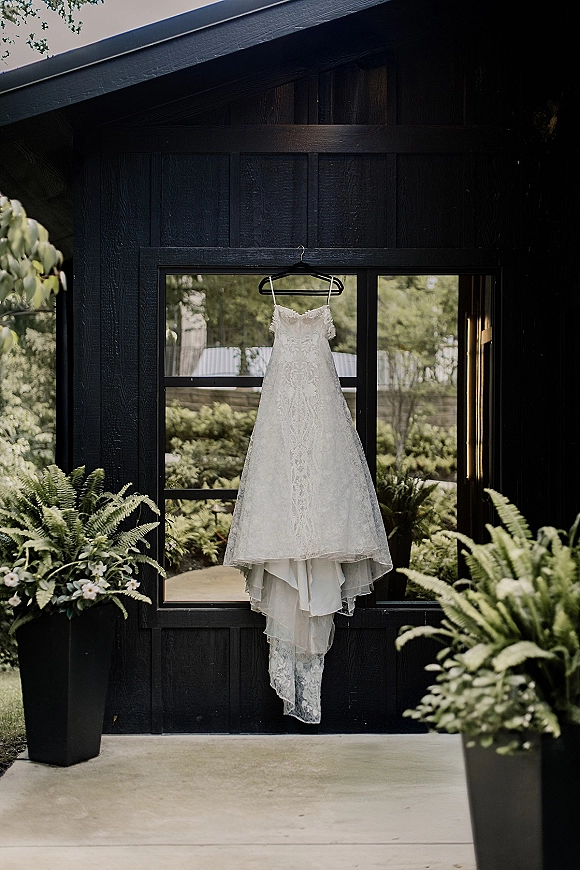 Wedding dress lace wedding dress hanging on a hanger by a window, framed by potted ferns and white flowers against dark wood siding