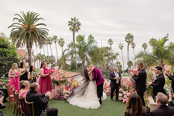 Ceremony kiss as bride and groom kiss at the altar, her cathedral veil flowing, with palm trees and ocean view behind guests and florals