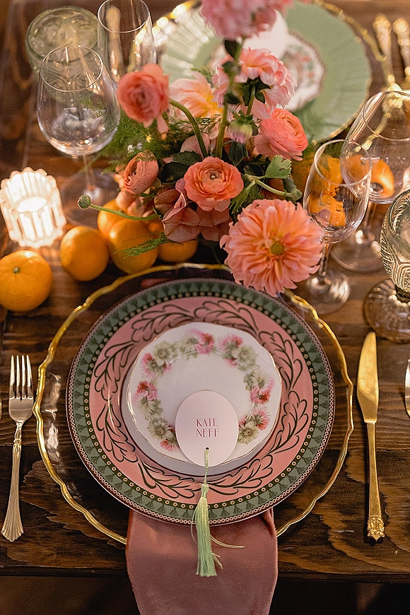 Reception tablescape with wedding place setting, pink floral centerpiece, oranges, taper candles, vintage plates, and gold flatware on a wood table